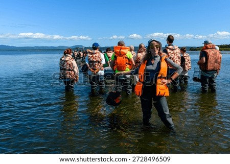 Groups of enthusiastic students wade through shallow water, observing nature while wearing life jackets and waiting to learn. The bright sun enhances the vibrant landscape.