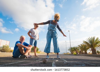 Grouyp Of Threee People Of Different Ages Playing Together At The Hopscotch On The Asphalt Of The Street - Seniors And Adult Woman Having Fun