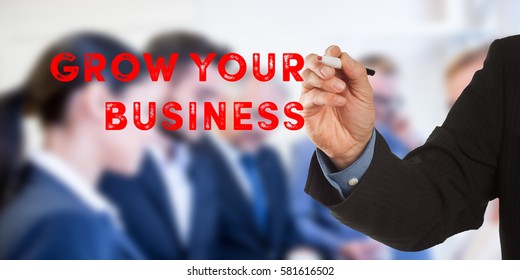 Grow your business, Male hand in business wear holding a thick pen, writing on an imaginary screen at the camera, business team in background, digital composing.