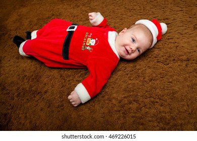 A Growing Baby Boy Celebrates His First Christmas In A Sleeper And Santa Hat That Are Too Small For Him.  He Is Laying On The Carpet And Smiling For The Camera.