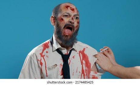 Growling scary zombie getting vaccine imunisation shot on blue background. Frightening brain-eating creepy monster with deep and bloody scars and wounds getting flu shot injection. Studio shot