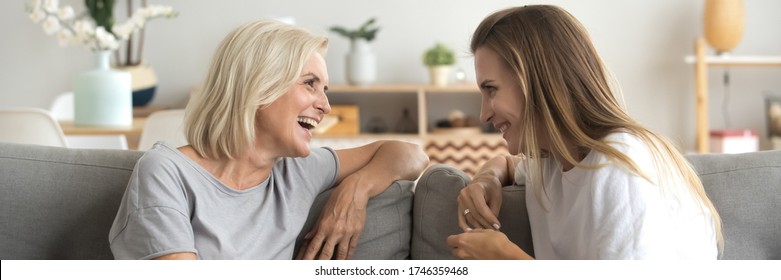 Grown up adult daughter middle-aged 60s mother seated on sofa having fun, two women gossiping enjoy positive conversation spend time together at home. Horizontal photo banner for website header design