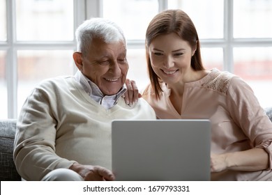 Grown up daughter and 80s father sit on couch with laptop. Nurse caregiver spend time with mature patient watching funny movies for positive mood, older younger relatives make videocall in living room - Powered by Shutterstock