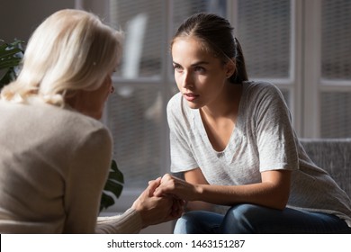 Grown up daughter holds hands of mid aged mother sit on armchair looking at each other having heart-to-heart talk, concept of close relative person, diverse generations connection, mom and adult child