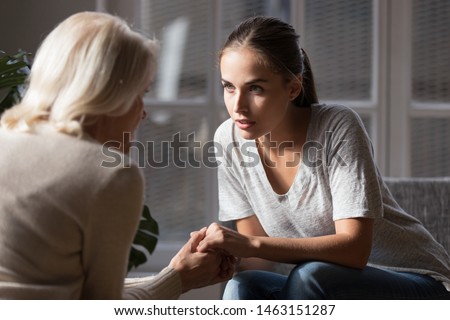 Grown up daughter holds hands of mid aged mother sit on armchair looking at each other having heart-to-heart talk, concept of close relative person, diverse generations connection, mom and adult child