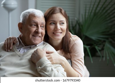 Grown Up Loving Attentive Caring Granddaughter Hugging Elderly 75s Grandfather. Young Adult Daughter Embracing Aged Father Looking At Distance In Cozy Living Room. Concept Of Family Ties Love And Care