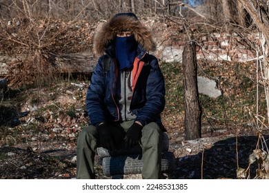 Un hombre adulto con una máscara, con una chaqueta encapuchada y con pantalones militares engullidos se sienta y mira a la cámara