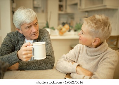 Grumpy Annoyed Caucasian Man On Retirement Sitting At Kitchen Table With Cup Of Coffee Arguing With His Attractive Wife, Having Disagreement About Plans. People, Aged, Relationships And Domestic Life