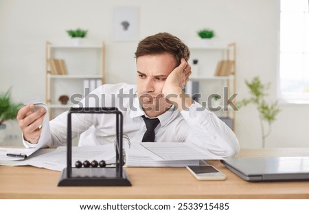 Grumpy bored overworked businessman looking unhappily at stack of paper work, sitting at work desk with Newton's cradle pendulum at foreground, having no motivation. Burnout and overwork concept.