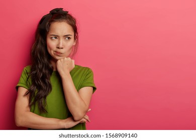 Grumpy displeased Asian female smirks face, keeps hands half crossed, holds chin, has angry expression, being deep in thoughts, dressed in casual wear, models over rosy wall, copy space aside