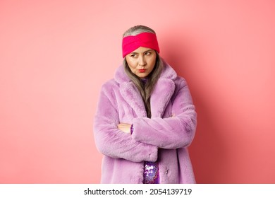 Grumpy Funky Old Asian Lady Looking From Under Forehead, Pouting And Looking Left With Angry And Disappointed Face, Standing In Stylish Purple Faux Fur Coat Against Pink Background