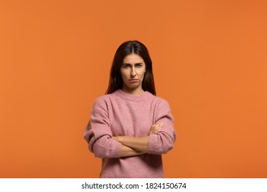 Grumpy irritated looks unhappy. Portrait of serious woman in pink sweater, has serious confident expression, keeps arms crossed, feels angry after quarrel with somebody. isolated on orange wall
