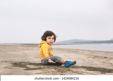 Grumpy Looking Two Years Old Toddler Boy Sitting At The Beach, Wales, United Kingdom.