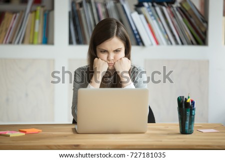 Grumpy not amused office worker looking at laptop screen in office. Young business lady bored with mundane workflow, tired from working too much. Casual businesswoman not happy with boring work task. 