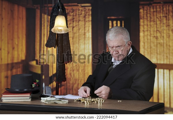 A grumpy old miser sitting at his desk\
counting gold coins by a stack of big\
bills.