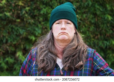 Grumpy Old Woman Stands Outside Against A Background Of Trees, With Long Hair, Wearing A Green Beanie And Plaid Jacket, Frowning                               