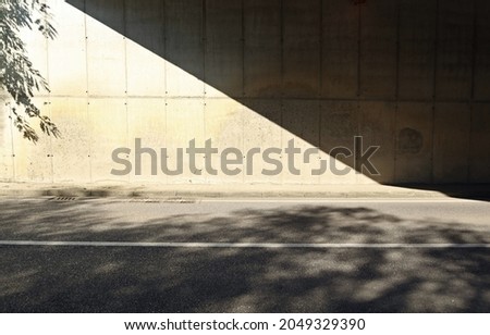 Grunge concrete wall of an underpass divided in two by the shadow of the bridge. Sidewalk and two lane road in front. Background for copy space.