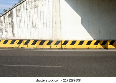 Grunge concrete wall of an underpass with a yellow and black guard rail divided in two by the shadow of the bridge Asphalt road in front. Background for copy space. - Shutterstock ID 2191299809