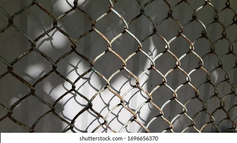 Grunge Rusty Wire Mesh Texture Closeup. Metal Lattice Pattern With Shadows In Bright Sunlight. Old Distressed Chainlink Fence In Front Of Grey Wall Background With Blurry Shadows.