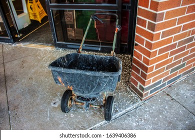 Grungy Two Wheeled Plastic Salt Spreader Parked Outside A Store Sliding Glass Door With Salt Scattered On The Sidewalk Around It After A Snow