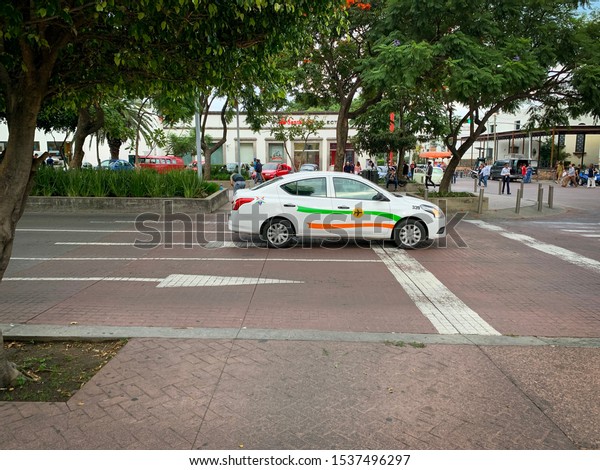Guadalajara, Mexico - October 19 2019: Sideview\
of white airport taxi stops at traffic light on its way to the\
Guadalajara\
airport