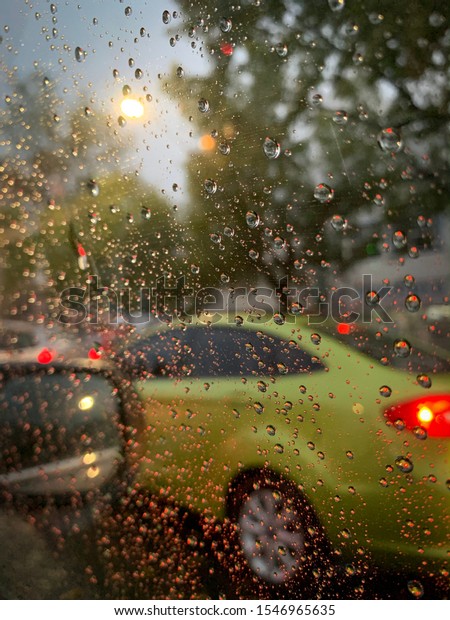 Guadalajara, Mexico - October 31 2019:\
Raindrops on car window during heavy rain. Green car in traffic,\
green trees with red highlight\
reflection