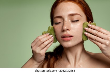 Guasha face massage with jade stone. Young redhead woman with freckles, massaging facial skin with gua-sha beauty scrapper, self-massage, spa procedure at home, green background