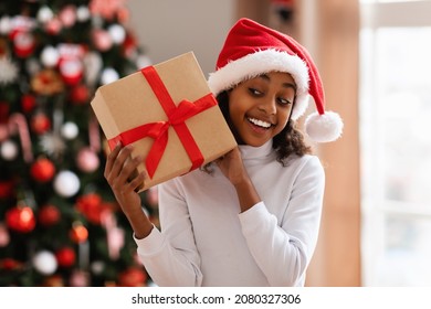 Guess What's Inside. Closeup Portrait Of Happy Girl Wearing Red Winter Santa Claus Hat Listening What Rattles In Gift Box. Curious Kid Shaking Christmas Present, Celebrating In Decorated Living Room