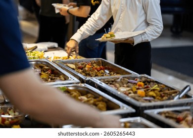 Guests are enjoying a lavish buffet with an array of colorful dishes, including meats, vegetables, and desserts, at a catered event in a banquet hall during the evening.