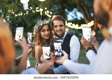 Guests with smartphones taking photo of bride and groom at wedding reception outside. - Powered by Shutterstock