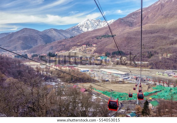 Guests of Sochi Winter Olympics rise by a\
cable way lift to RusSki Gorki Ski Jumping Center to watch the\
competitions. Esto-Sadok railway hub on the background. Blue sky\
mountain landscape.