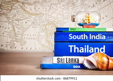 Guide books on the table with Seashell and ship in the bottle on its at old map background. Books with titles: South India, Bali, Sri Lanka, Goa, Thailand - Shutterstock ID 86879911