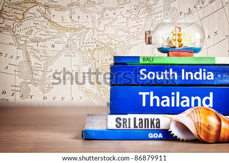Guide books on the table with Seashell and ship in the bottle on its at old map background. Books with titles: South India, Bali, Sri Lanka, Goa, Thailand