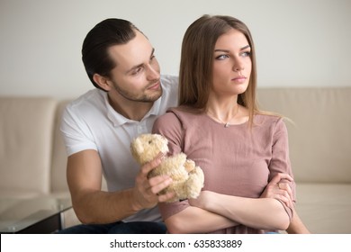 Guilty boyfriend asking for forgiveness, presenting offended girlfriend a teddy bear toy, lady looking proud sitting with arms crossed not going to take gift. Reclaim a fault, apology not accepted