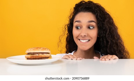 Guilty Pleasure Concept. Funny Hungry Woman Looking At Burger On Plate, Peeking Out Table Biting Lips, Excited Lady Craving Tasty Sandwich Isolated On Yellow Orange Studio Background. Panorama Banner