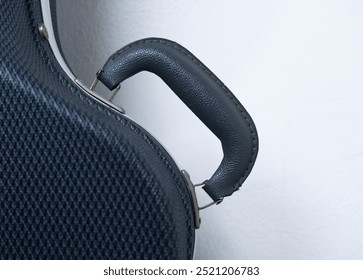 Guitar case handle with white wall background. Close-up dark grey leather of handle for guitar. Concept of fibers, box, plastic, classic, and vintage. Hard case of instrument for protection on wall.