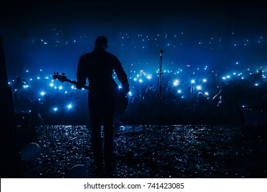 Guitar Player In Front Of Crowd On Scene In Night Club. Bright Stage Lighting, Crowded Dance Floor. Phone Lights At Concert. Band Blue Silhouette Crowd. People With Cell Phone Lights.