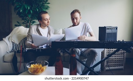 Guitarist And Keyboarder Are Talking Looking At Music Sheet And Discussing Song Lyrics During Rehearsal Break. Electric Musical Instruments, Drinks And Snacks Are Visible.