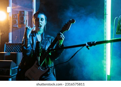 Guitarist woman standing in front on microphone while playing at electricguitar working at rock album in studio. Musician with rockstar outfit preparing heavy metal song for grunge performance
