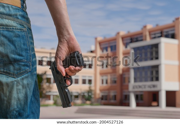 Gun control concept. Young armed\
man holds pistol in hand in public place near high\
school.