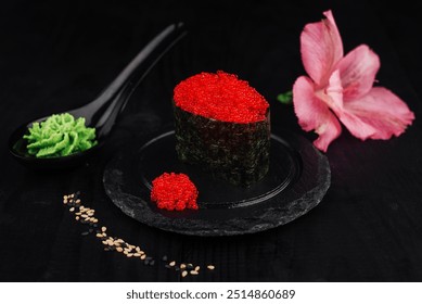 Gunkan maki sushi with tobiko flying fish roe is presented on a black slate plate with wasabi and a pink flower - Powered by Shutterstock