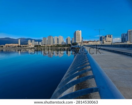 Gurney Bridge, an important link between Gurney Drive, and the small town in the Andaman Islands. City skyline reflects in calm waters below. Modern buildings stretch behind gentle clouds. Walking path invites quiet strolls beside metal railings.