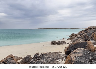 Gurteen Beach at sunset showcases clear turquoise waters and a rocky coastline in Ireland. The soft white sand contrasts with dark, weathered rocks under a moody overcast sky - Powered by Shutterstock