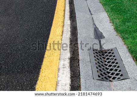 gutter of a stormwater drainage system on the side of an road with yellow and white markings and green lawn on wet weather after rain, close up view, nobody.