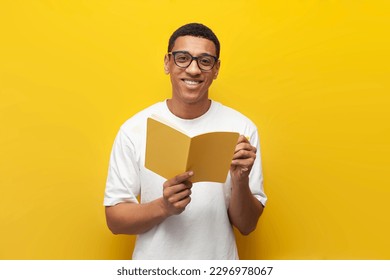 guy african american student in white t-shirt and glasses writes in notebook and reads book on yellow isolated background, smart man holds book - Powered by Shutterstock