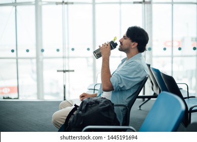 The Guy At The Airport Waiting For Departure, Drinking Water. A Young Man Waits For Boarding A Plane, Sits With A Backpack At The Gate, Drinks Water From A Bottle