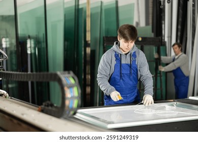 Guy apprentice in blue overall on cutting table trims and aligns edges of workpiece, creating plastic sheathing sheets.