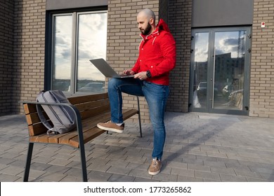 A Guy With A Beard In A Red Jacket And Jeans Is Working At A Computer On The Street. The Concept Of Freelance Work