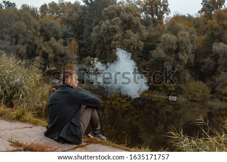 A guy with a beard sits on the lake and smokes an electronic cigarette. Blows puffs of smoke. The concept of unity with nature. Rest near the water. Vape, Man Vaping.