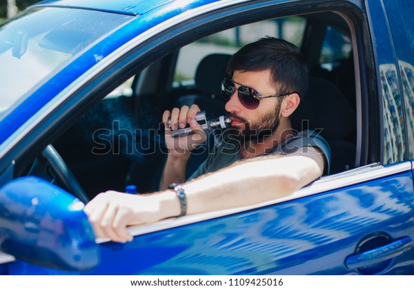 A guy with a beard in sunglasses is\
sitting in the car and smokes an electronic\
cigarette